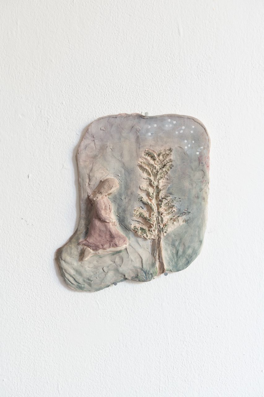 Wandrelief aus Ton in unregelmäßiger Form: eine kniende Figur neben einem zarten Baum, in Grau-, Rosa- und Grüntönen