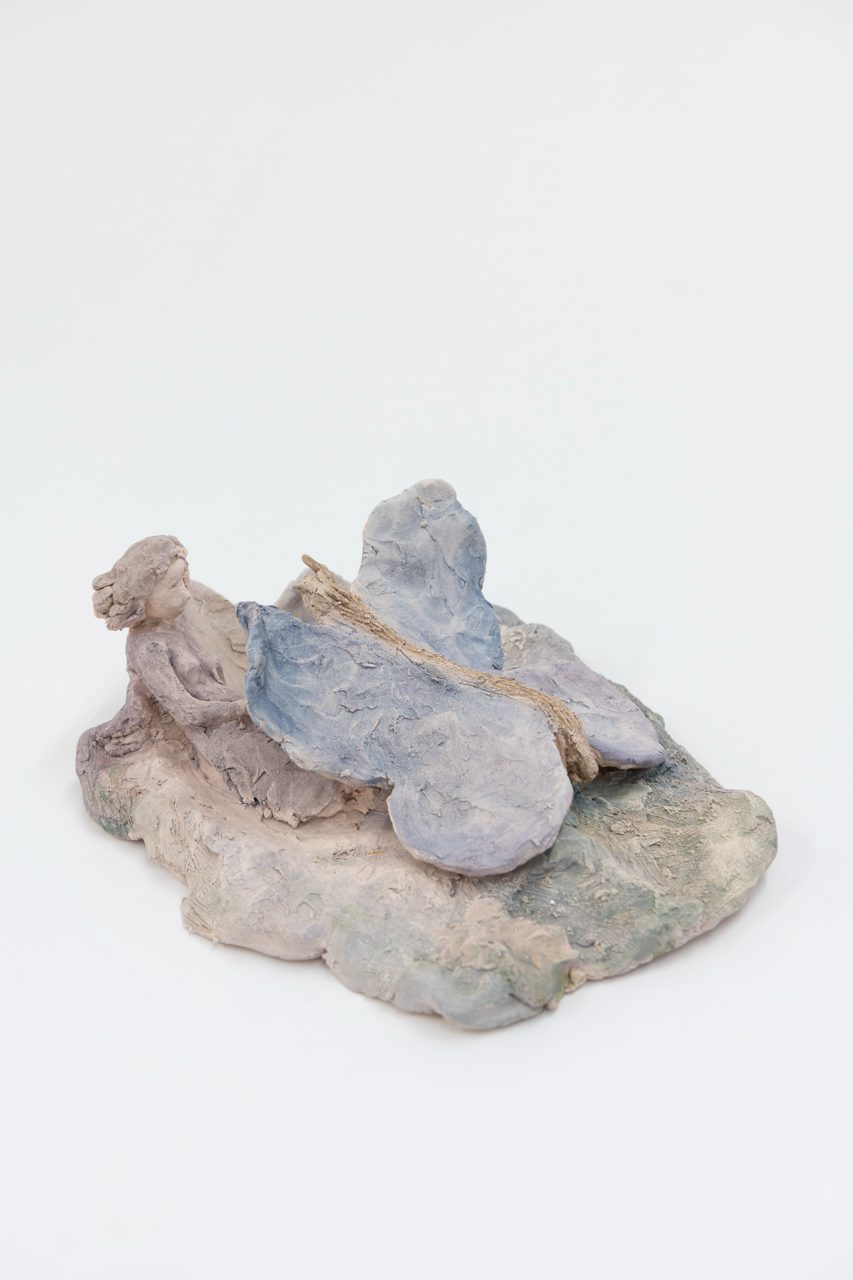 Liegende Mädchenfigur aus Ton mit großem blauem Schmetterling auf einer moosgrünen Platte